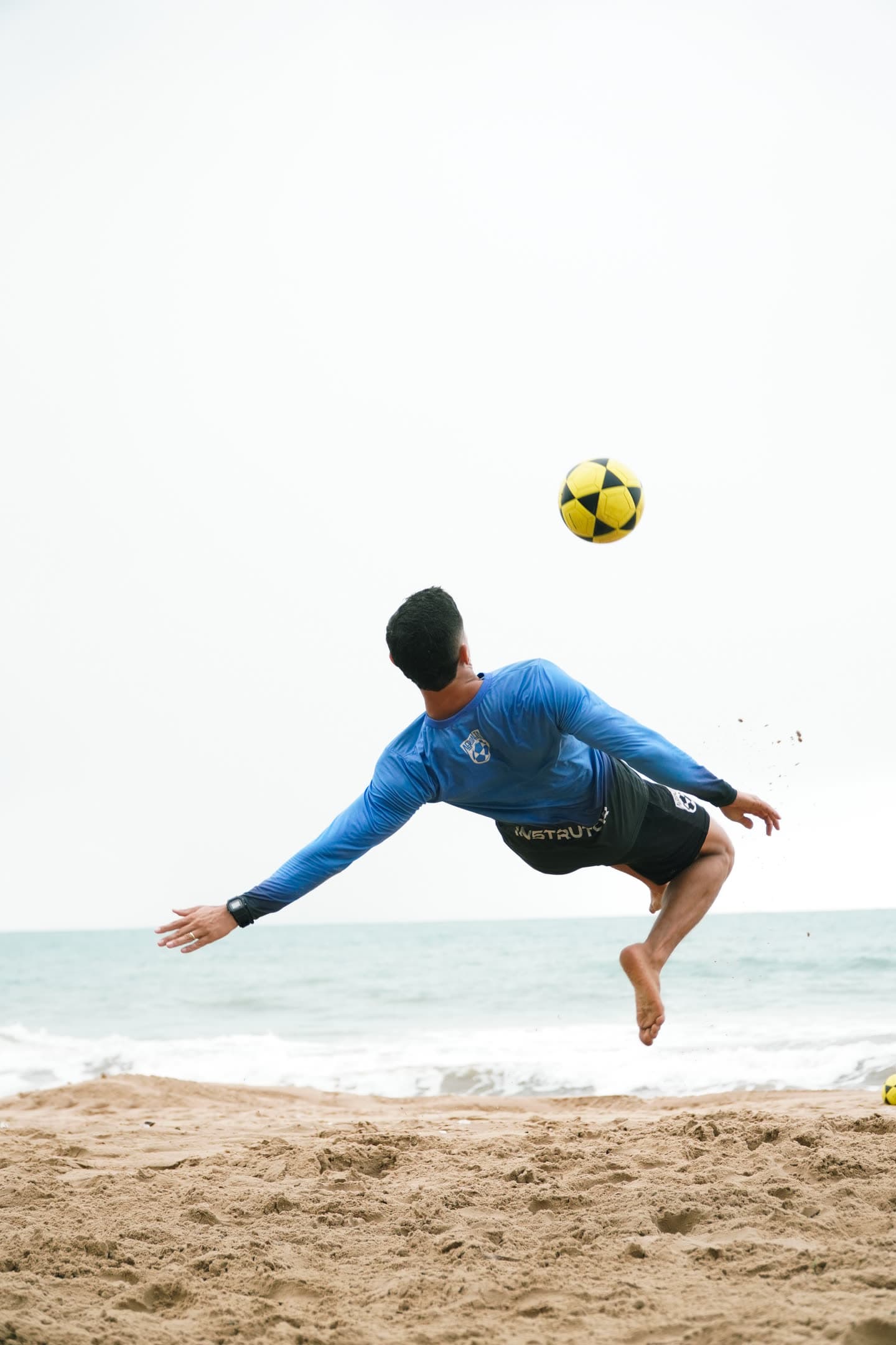 Atleta de camisa azul girando em bicicleta na areia, com a bola no ar e o mar ao fundo