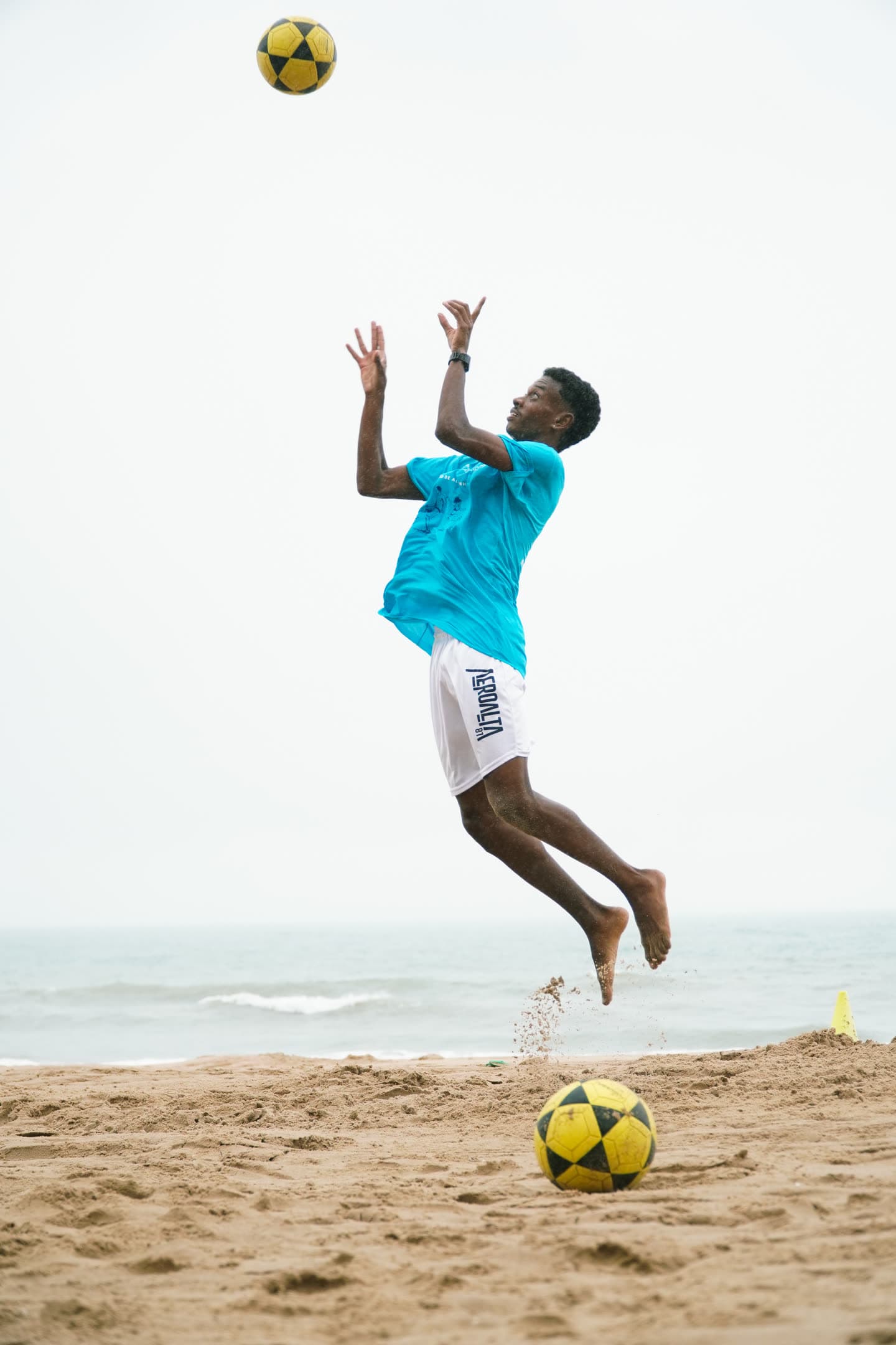 Atleta de camisa turquesa saltando com os braços erguidos para alcançar a bola, na beira do mar