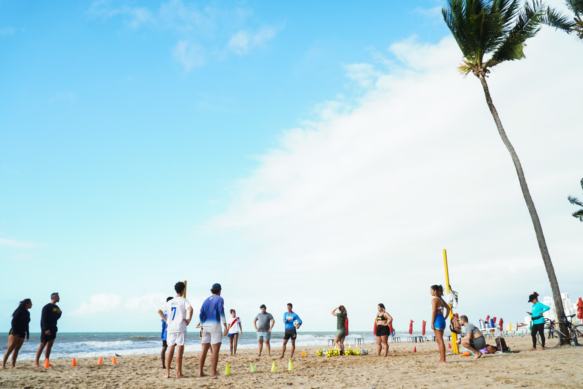 Alunos em fila durante treino de altinha na beira do mar, com cones e bolas na areia