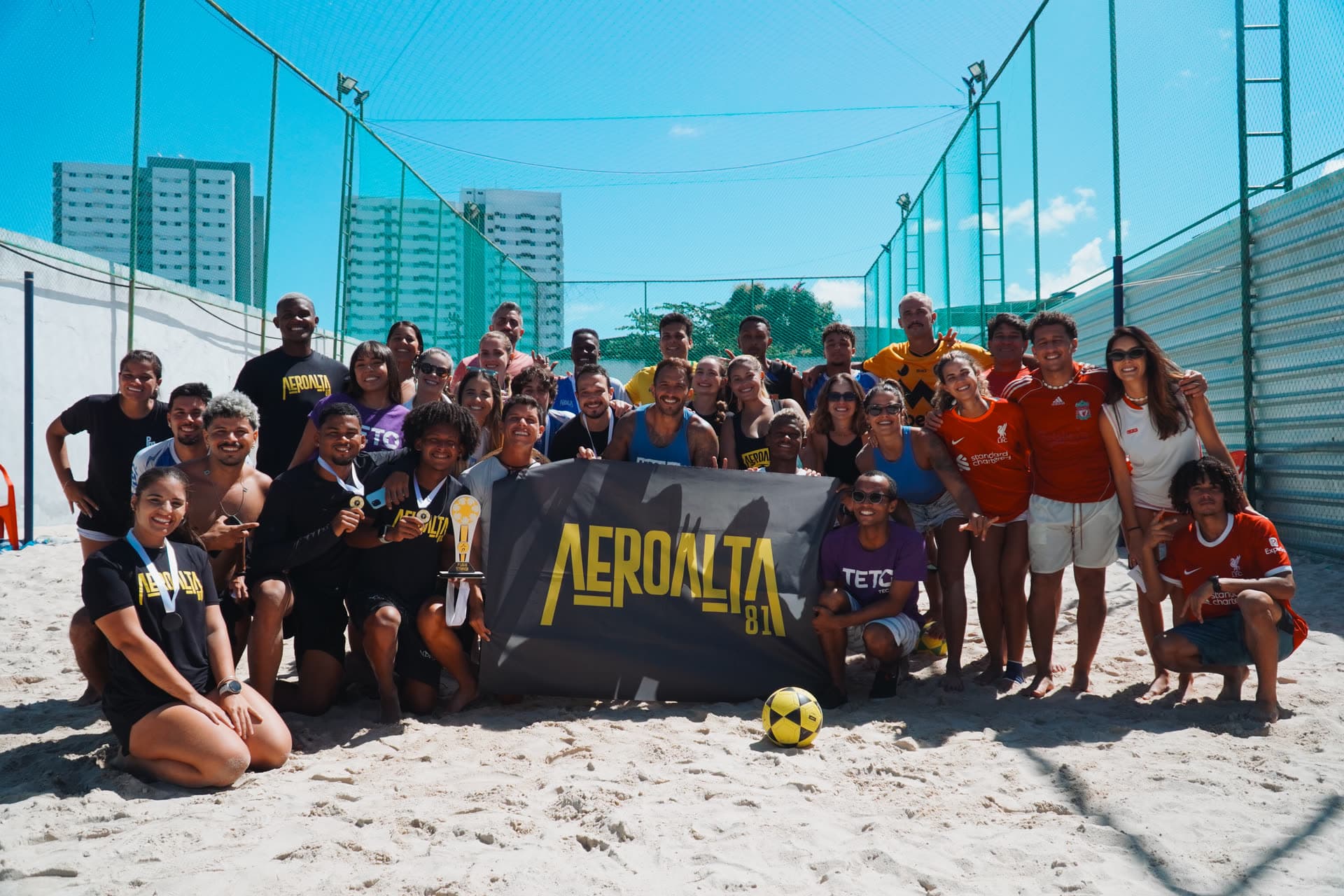 Turma do Aero Alta 81 posando com a bandeira da liga e o troféu na quadra de areia