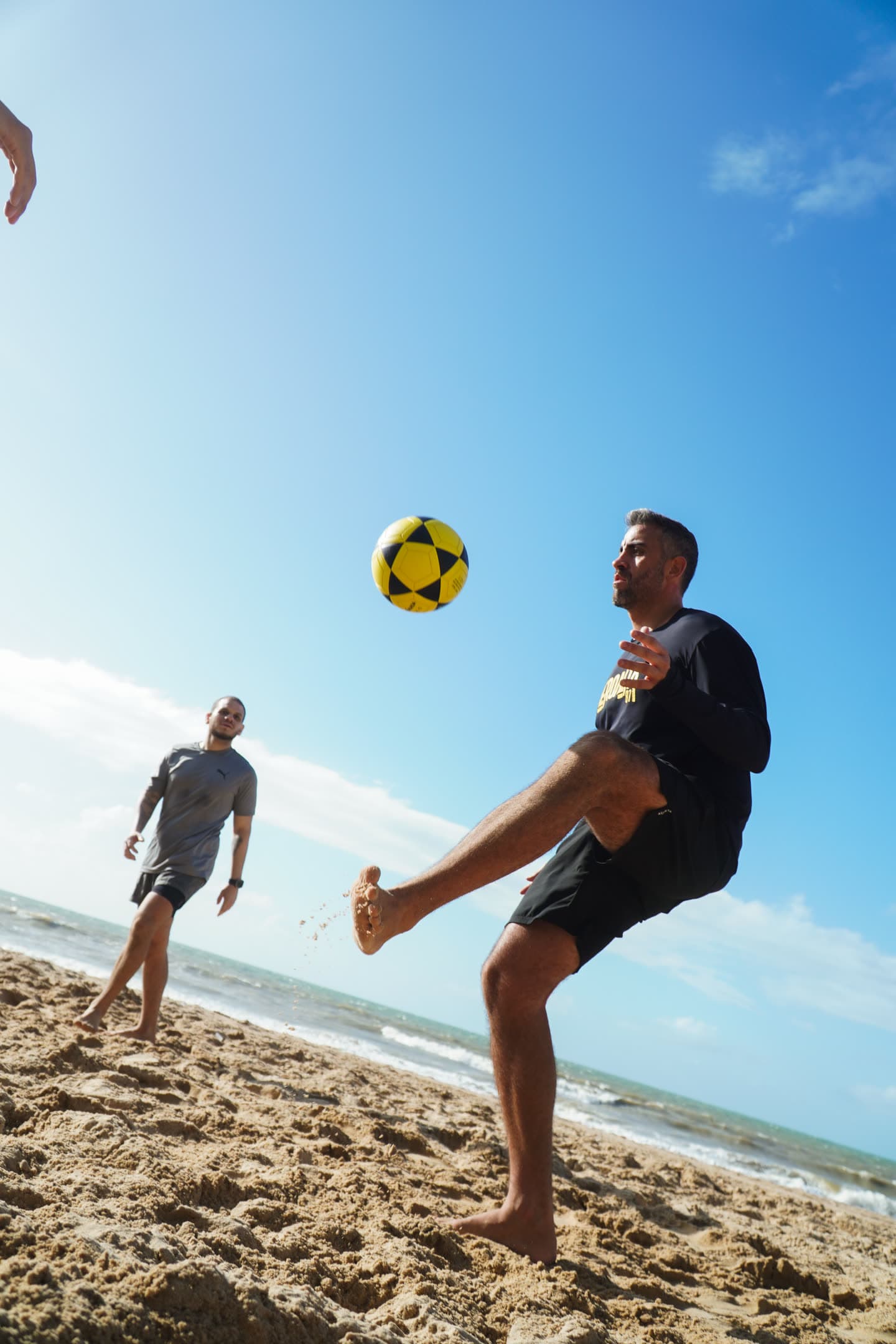 Atleta de preto erguendo a perna para o voleio, com a bola amarela e preta contra o céu